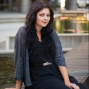 A woman with long black hair and wearing a black dress and gray shawl sits on a wooden dock in front of a body of water.