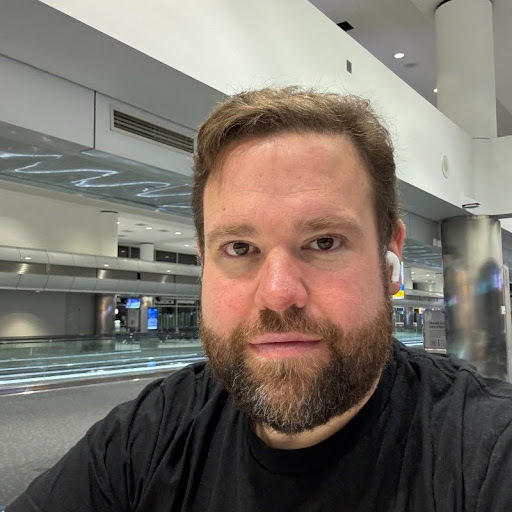 A bearded man wearing a black shirt and wireless earbuds sits in a brightly lit, modern airport terminal.