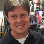 A person with short brown hair smiles at the camera. Behind them are various posters, chairs, and equipment.